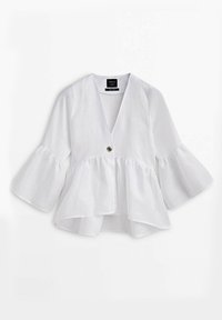 Blouse blanche à manches longues avec col en V, bouton central unique, poignets volantés évasés et ourlet péplum froncé.