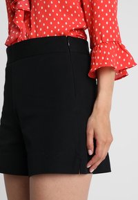 Pantaloni corti neri con una superficie liscia, dotati di una vita aderente e fessure laterali. Indossati con una blusa rossa decorata con pois bianchi.