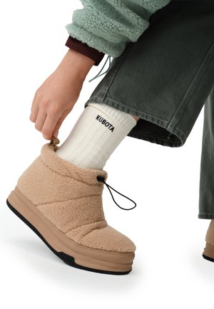 Beżowe pluszowe kapcie o wysokim kroju z teksturowanym wykończeniem i czarną gumową podeszwą. Posiadają ściągacz i zaokrąglony czubek.