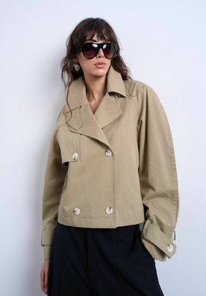 Kvinde iført oversized beige dobbeltradet jakke med bred krave, opkørte ærmer, sorte bukser, store solbriller og øreringe i hoops mod hvid væg.