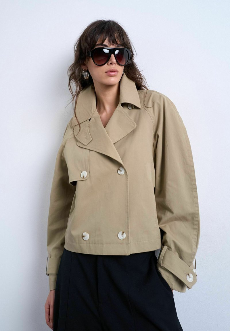 Mujer con chaqueta beige de gran tamaño, cruzada y con cuello ancho, mangas remangadas, pantalones negros, grandes gafas de sol y pendientes de aro, posando frente a una pared blanca.