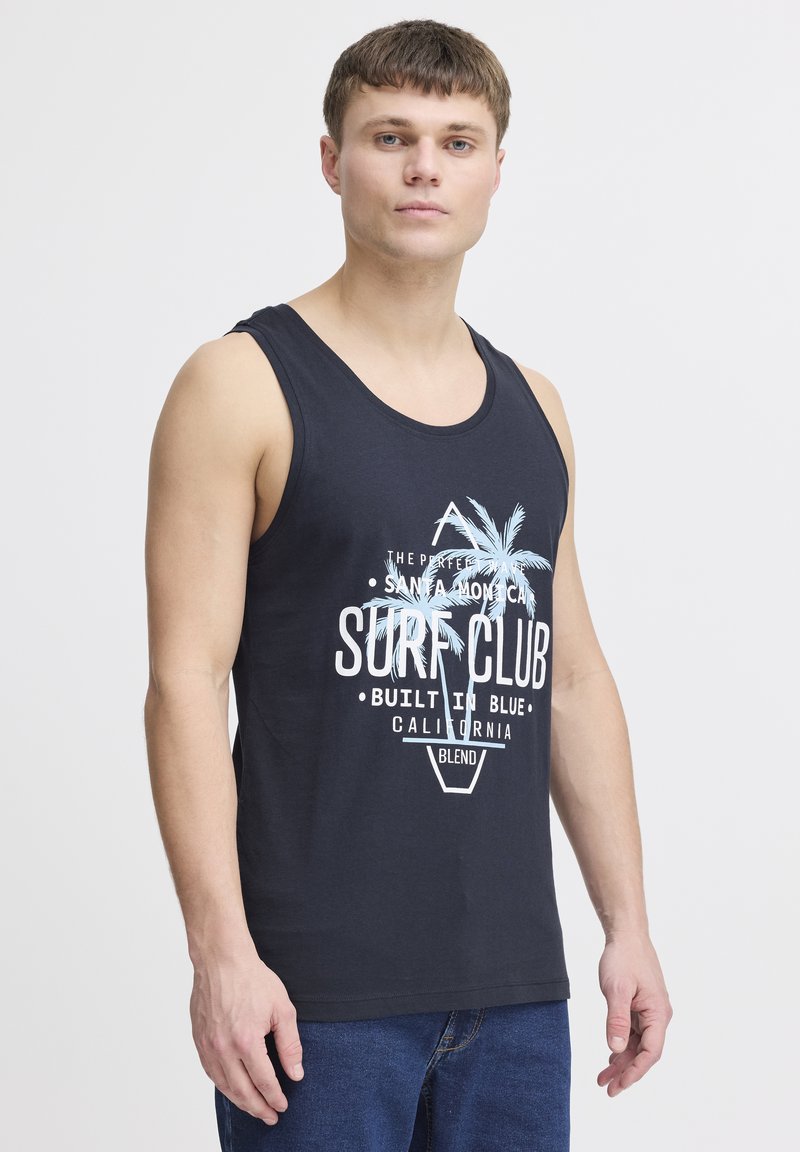 Černý top bez rukávů s bílým grafickým potiskem, který zobrazuje palm trees a text. Měkká látka, volný střih. Nosený s denimovými džíny.