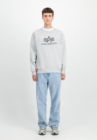 Felpa grigia con logo, con polsini e orlo a costine, abbinata a jeans azzurri e sneakers grigie. Modello in piedi su uno sfondo semplice.