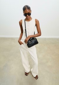 Sac en cuir noir avec quincaillerie dorée, associé à un top sans manches crème et un pantalon large crème. Le modèle porte des lunettes de soleil noires surdimensionnées.