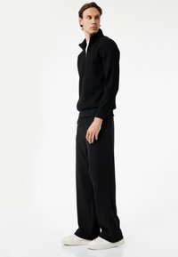 Sort lynlåstrøje med et tekstureret stof, parret med løse sorte ribbede bukser og hvide sneakers, modelleret af en mandlig figur.