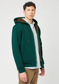 Felpa verde con zip e fodera in pile marrone, con coulisse regolabili e tasche frontali. Indossata sopra una camicia a quadri azzurri.