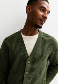 Cardigan verde lavorato a maglia con un motivo texture, scollo a V e cinque bottoni. Indossato sopra una maglietta di colore chiaro con una texture liscia.