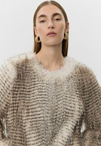 Lažni krzneni pulover s prugastom teksturom, koji ima široki ovratnik i mekan, pahuljast materijal u nijansama smeđe i kreme.