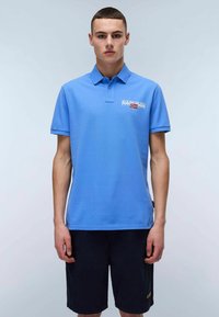 Chemise polo bleue en coton, avec un col, des manches courtes et un logo imprimé sur la poitrine gauche. Portée avec un short bleu marine.