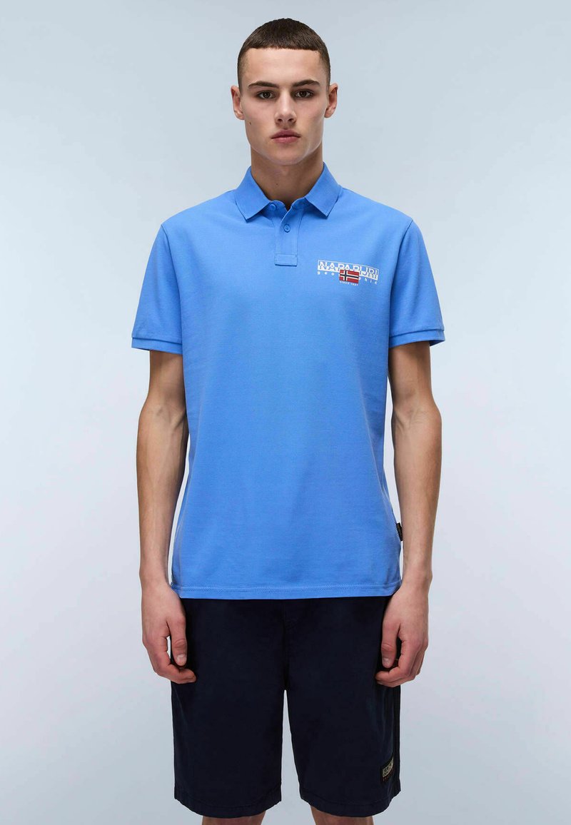Chemise polo bleue en coton, avec un col, des manches courtes et un logo imprimé sur la poitrine gauche. Portée avec un short bleu marine.