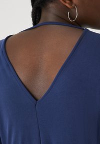 Mornarsko modra majica z obliko V na hrbtnem delu. Gladka tekstura tkanine z vertikalno šivano podrobnostjo na sredini. Enostaven, minimalističen slog.