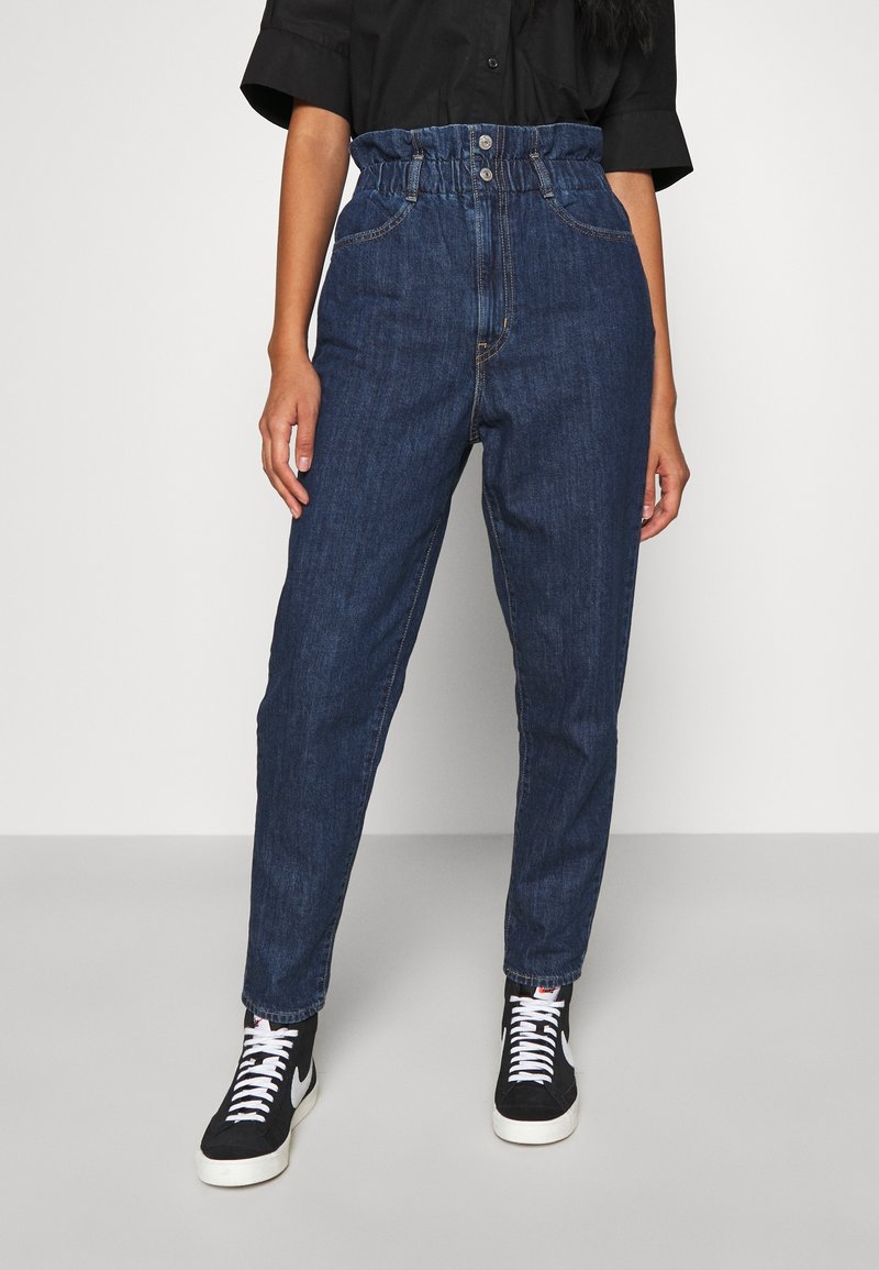 Personne portant un jean bleu taille haute avec une ceinture paperbag, un t-shirt noir et des baskets montantes blanches et noires, debout sur un sol uni.