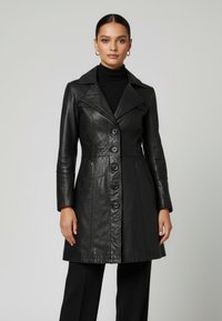 Kvinna som bär en figursydd svart läderrock med knappar över svarta byxor och polotröja, stående mot en enkel ljus bakgrund.