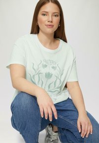 Lichtgroen katoenen T-shirt met korte mouwen, voorzien van een plantenthema grafisch ontwerp met het woord "GRATEFUL." Gecombineerd met blauwe denim jeans.