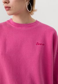 Ljust rosa sweatshirt med ribbad rund hals och subtil broderad logotypdetalj på bröstet. Mjuk tyg med avslappnad passform.
