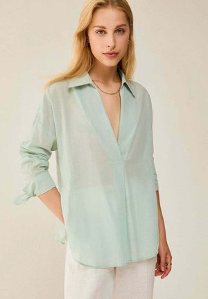 Donna bionda con trucco leggero che indossa una camicia larga verde pallido senza bottoni e pantaloni bianchi, in piedi con una mano in tasca.