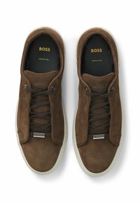 Ein Paar braune Wildleder-Sneaker von BOSS mit dunklen Schnürsenkeln, weißen Sohlen und markierten metallenen Schnürsenkelanhängern, von oben auf einem weißen Hintergrund betrachtet.