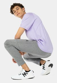 Ein helllila Sport-T-Shirt, graue gemusterte Jogginghosen und schwarz-weiße Turnschuhe. Die Jogginghosen haben eine enganliegende Passform und Knöchelbündchen.