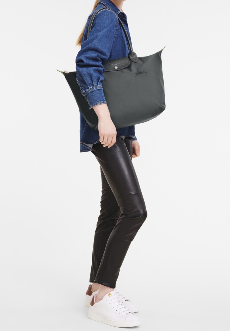 Personne portant une veste en jean bleue, un pantalon en cuir noir, des baskets blanches et portant un grand sac à main noir sur l'épaule.