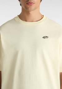 Cremefarbene Baumwoll-T-Shirt mit Rundhalsausschnitt und kurzen Ärmeln. Verfügt über ein kleines schwarzes Vans-Logo auf der linken Brustseite.
