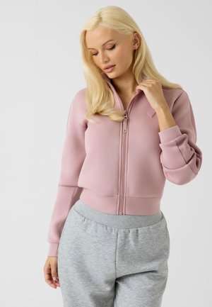 Femme aux longs cheveux blonds portant une veste rose zippée et un pantalon de jogging gris clair, regardant vers le bas avec une main près du col.