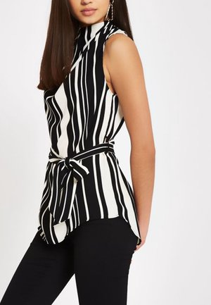 Kvinde iført sort og hvid stribet top uden ærmer med knude i taljen, kombineret med sorte tætsiddende bukser, står mod en hvid baggrund.