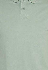 Světle zelená polo košile s límečkem, se dvěma knoflíky u krku, vyrobená z texturované látky. Jednoduchý, klasický design.
