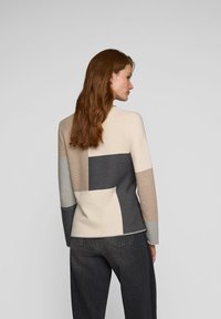 Gestrickter Pullover in Beigetönen, Grau und Taupe-Patches. Mit Rundhalsausschnitt und langen Ärmeln. Kombiniert mit weiten dunkelgrauen Jeans. Glatte Textur.