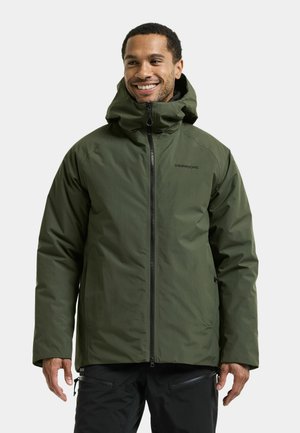 Man smiling wearing an olive green hooded zip-up winter jacket and black pants against a plain light background.