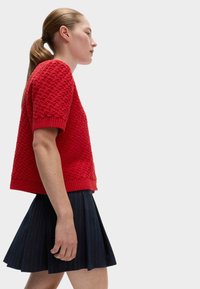 Czerwony sweter dziany w krótkie rękawy z teksturowanym wzorem, w połączeniu z ciemną spódnicą plisowaną z cienkimi pionowymi paskami.