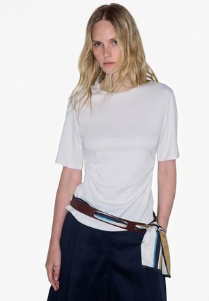 Donna bionda che indossa una semplice maglietta bianca a maniche corte, pantaloni blu scuro e una cintura marrone con una sciarpa multicolore annodata come accessorio.