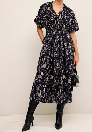 Marineblaues Kleid mit abstraktem Multicolor-Muster, kurzen Puffärmeln, taillierter Taille und fließendem Rock. Kombiniert mit hohen schwarzen Stiefeln mit Absatz und einer kleinen schwarzen Tasche.