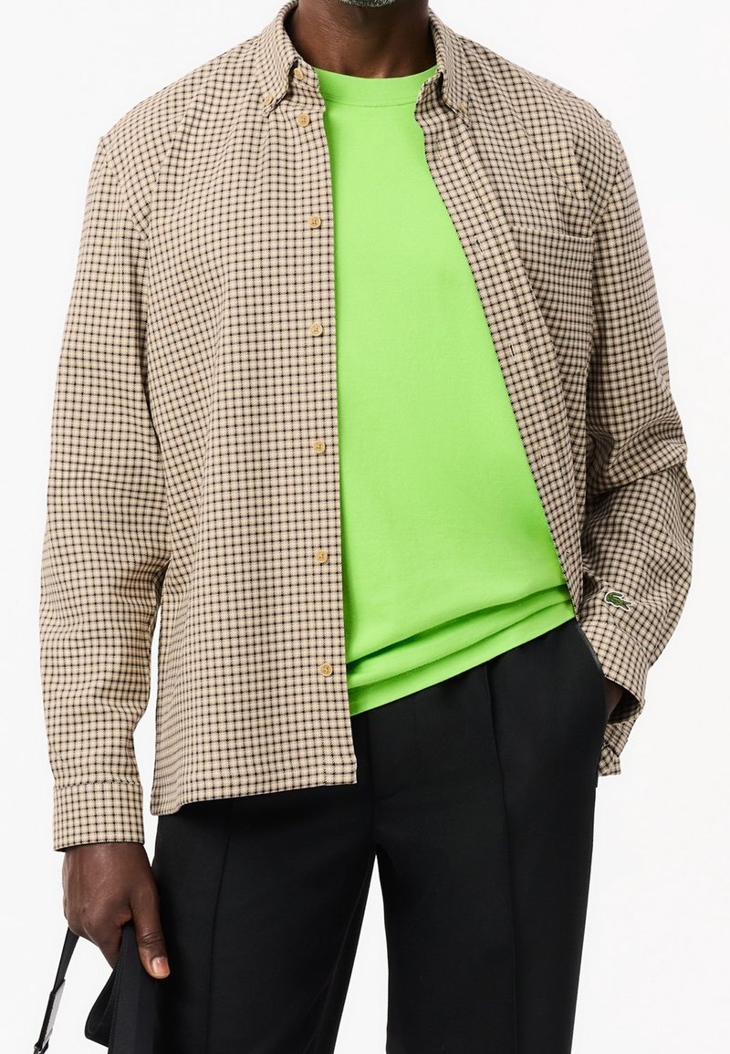 Brązowa koszula w kratę z guzikami i kołnierzykiem, nałożona na jasnobrązowy t-shirt. Czarne spodnie dopełniają stylizację, eksponując kontrastujące kolory.