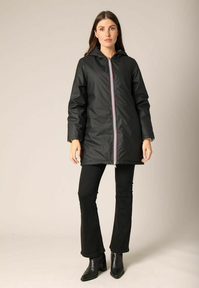 Veste imperméable noire avec capuche, dotée d'une fermeture éclair frontale complète et de détails à rayures colorées. Portée avec un pantalon noir évasé et des bottines à talons.