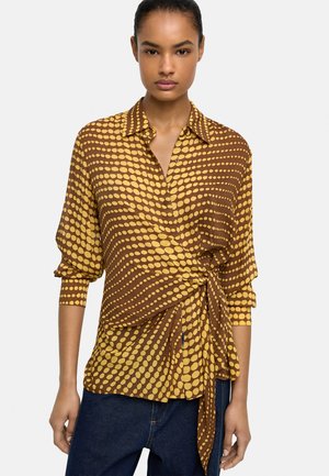 Kobieta ubrana w brązową i żółtą bluzkę z długimi rękawami w groszki, do tego ciemnoniebieskie jeansy, stojąca na jednolitym tle.