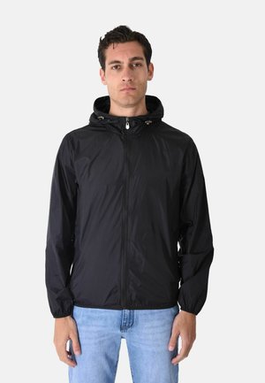 Uomo di fronte che indossa una giacca nera con cappuccio e zip, polsini elastici e jeans azzurri su uno sfondo uniforme.