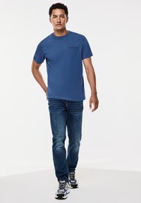 Sininen puuvillapaita lyhythihaisena ja pienellä mustalla logolla rinnassa, yhdistettynä sinisiin farkkuihin ja urheilullisiin mustiin kengiin.