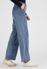 Pantalons en velours côtelé à jambes larges de couleur bleu clair avec des rayures verticales, dotés de poches latérales, associés à des baskets beiges et dorées.