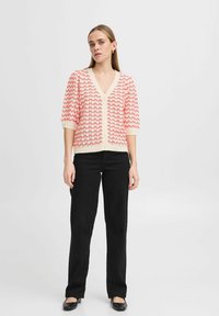Un cardigan rose et crème avec un col en V, des manches courtes et un motif ondulé texturé ; complété par une fermeture à boutons et porté sur un pantalon noir.