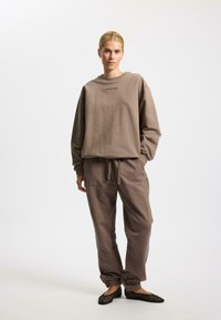 Sudadera marrón y pantalones a juego de tela suave, con un corte relajado, puños acanalados y cintura con cordón. Detalle en texto negro en la sudadera.