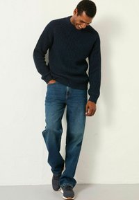 Marineblauer gerippter Strickpullover mit langen Ärmeln, kombiniert mit blauen Jeans und dunkelgrauen Schuhen. Das Model steht lächelnd vor einer hellen Wand.