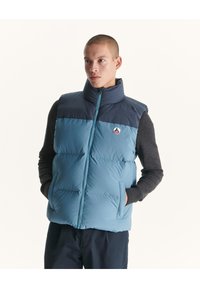 Lichtblauw puffer vest met een hoge kraag, marineblauwe bovenkant en zijzakken. Voorzien van een rits aan de voorkant en een logo badge op de borst.