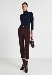 Maglione a collo alto testurizzato blu navy, pantaloni borgogna su misura con cintura e stivali alla caviglia lucidi color borgogna. Porta una piccola borsa a mano scura.