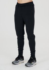 Sorte atletiske joggers med snøre i taljen, taperede ben og lynlås sidelommer, kombineret med sorte og grå løbesko.