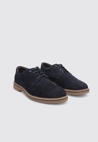 Zapatos de ante azul marino con cordones, puntera redondeada, detalles en piel y suela de goma color marrón claro en contraste. Textura suave y diseño clásico.