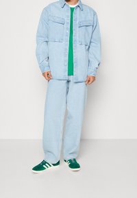 Light blue denim shirt with two chest pockets, green t-shirt underneath, light blue denim pants, and green suede sneakers with white stripes.