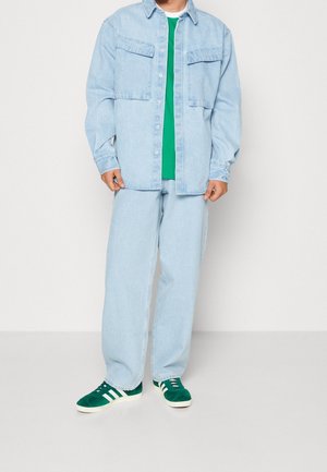 Chemise en denim bleu clair avec deux poches poitrine, t-shirt vert en dessous, pantalon en denim bleu clair et baskets en suède vert avec des rayures blanches.