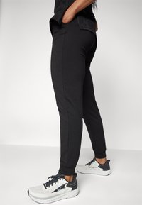 Mustad vööstatud jooksutraksid kitsas lõikega, elastsete mansettide ja taskutega, millele on kombineeritud hallid ja valged spordijalatsid, millel on tekstuuriga disain.