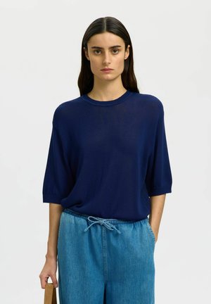 Kobieta o długich ciemnych włosach nosi granatowy sweter z krótkim rękawem i luźne niebieskie dżinsy z sznurkiem w talii, stojąc na białym tle.