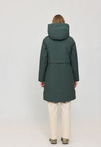 Lange grüne gepolsterte Jacke mit Kapuze, tailliertem Schnitt und seitlichen Taschen. Glattes, strukturiertes Material, das bis zur Mitte des Oberschenkels reicht. Kombination mit cremefarbener Hose.